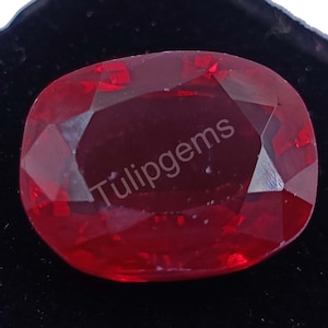 May include: A faceted, oval-shaped, deep red gemstone with the text "Tulipgems" in white. The gem has a high luster and is set against a dark background, highlighting its color and clarity. This gemstone is likely for jewelry.