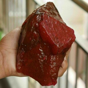 May include: A large, rough-cut, deep red gemstone held in a hand. The stone has a glossy, uneven surface with variations in colour and texture. The overall shape is irregular, with a prominent, flat plane on one side.