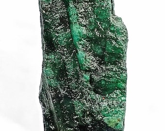 May include: A raw, uncut emerald gemstone. The deep green crystal has a rough, textured surface with visible natural formations. The gemstone is held against a dark fabric background, highlighting its vibrant color and natural beauty.