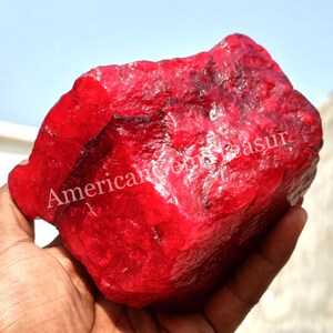 May include: A large, rough-cut ruby gemstone with a deep red color. The surface has a textured appearance, and the text "American Gemsasur" is visible on the stone. The gemstone is held in a hand against a bright sky.
