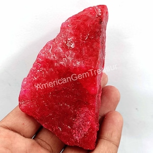 May include: A rough-cut, vibrant red gemstone with a textured surface. The stone has a natural, irregular shape and is held in a hand. The text "AmericanGemTreasure" is visible on the stone.