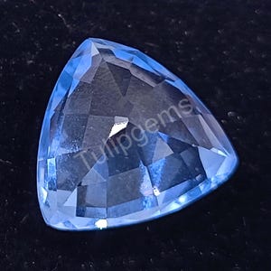 Puede incluir: Una gema triangular azul claro con múltiples facetas. La gema tiene una superficie pulida que refleja la luz. El texto "Tulipgems" es visible en la superficie. La gema está sobre un fondo oscuro.