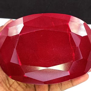 Könnte beinhalten: Ein großer, facettierter, ovaler Rubin-Edelstein. Der Edelstein hat eine tiefe, satte rote Farbe mit mehreren Facetten, die das Licht einfangen. Der Rubin wird in einer Hand gehalten, mit einem schwarzen Hintergrund.