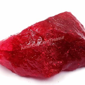 May include: A rough, uncut ruby gemstone in a deep red hue. The surface has a textured appearance. The text "American Gem Treasur" is visible on the surface of the gemstone.