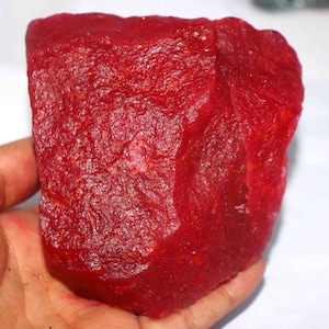 May include: A large, raw, uncut ruby gemstone with a deep, vibrant red color. The stone features a rough, crystalline texture with natural facets and irregular edges, showcasing a translucent quality when light hits the surface of the mineral.