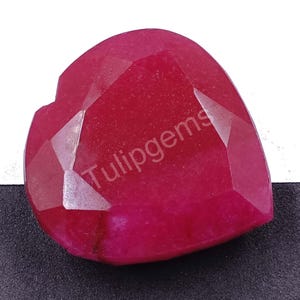 May include: A heart-shaped ruby gemstone with multiple facets. The gem is a deep red colour, with the text "Tulipgems" visible on the surface. The facets create a play of light across the surface of the stone.