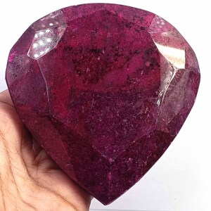 Op de afbeelding: Een grote, hartvormige edelsteen met een diepe magenta en donkerrode kleur. De steen heeft meerdere platte, gepolijste facetten over het oppervlak, wat zorgt voor een geometrisch uiterlijk met zichtbare interne texturen en reflecties.