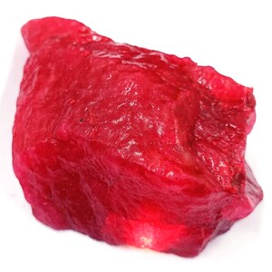 May include: A raw, uncut ruby gemstone with a deep red color. The surface has a rough, textured appearance, with variations in tone and highlights. The gemstone is set against a white background, emphasizing its natural form.