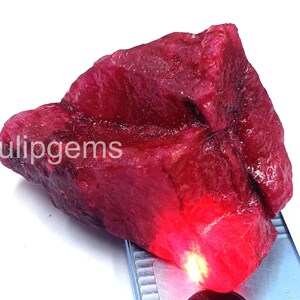 May include: A raw, uncut ruby gemstone in a deep red hue. The stone's surface has a textured appearance. The word "ulipgems" is printed on the stone. The gemstone is set against a white backdrop.