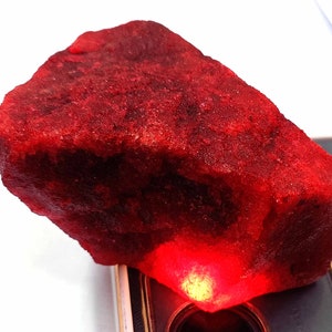 May include: A rough, uncut ruby gemstone with a deep red color. The surface of the stone has a textured appearance. The ruby is displayed on a reflective surface, with a bright light illuminating the bottom of the stone.