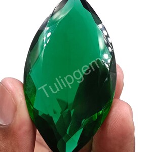 May include: A marquise-cut, faceted gemstone in a deep emerald green color. The gem is held in a hand and features the text "Tulipgem" in white. The gem's facets reflect light, creating a sparkling effect.