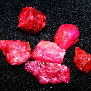 May include: A collection of raw, uncut ruby gemstones. The rubies display a vibrant red hue, with varying shapes and sizes. The rough texture and natural forms of the stones are visible against a dark background.