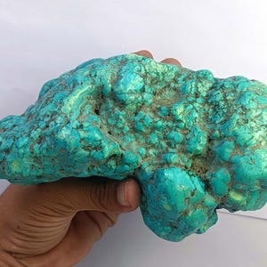 May include: A large, raw turquoise stone featuring a vibrant teal and blue colour palette with natural, irregular surface textures, deep crevices, and dark matrix veining throughout the mineral formation, showcasing its organic, unpolished appearance.