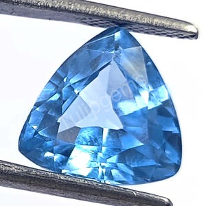 Puede incluir: Una gema facetada, triangular, de color azul claro, sujeta con pinzas metálicas. La gema tiene una superficie pulida con múltiples facetas que reflejan la luz. La gema es de un tono azul vibrante, con un aspecto claro. Imagen de primer plano.