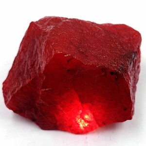 May include: A rough-cut, vibrant red gemstone with a textured surface. The stone has a natural, irregular shape and a glowing, internal light. The color is a deep, saturated red, with variations in tone across the surface.
