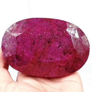 May include: A large, faceted ruby gemstone with a deep red hue. The oval-shaped stone has multiple facets that reflect light, creating a sparkling effect. The gemstone is held in a hand against a white background.