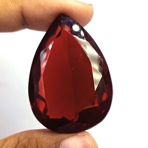 May include: A pear-shaped, faceted gemstone with a deep, translucent red colour. The stone features multiple triangular and trapezoidal facets across its surface, creating a reflective appearance with light highlights visible on the upper edges.