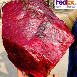 Puede incluir: Una gran gema de rubí en bruto, de color rojo intenso. La superficie es texturizada y presenta variaciones naturales. El logotipo de FedEx Express es visible en la esquina superior derecha.