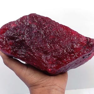 Puede incluir: Una gran gema de rubí en bruto, de color rojo intenso. La piedra en bruto se sostiene en una mano, mostrando su tamaño y textura. La superficie tiene una apariencia cristalina, reflejando la luz.