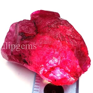 May include: A raw, uncut ruby gemstone with a vibrant red hue. The rough surface of the stone is visible, with areas of deep color and natural texture. The word "alipgems" is visible on the stone.