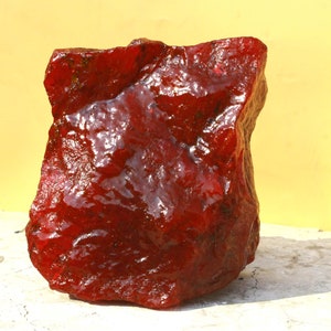 Puede incluir: Una gema grande, en bruto, de color rojo rubí con una superficie brillante y reflectante. La piedra tiene una forma irregular y un color profundo y saturado, lo que sugiere que es un espécimen natural sin cortar. El fondo es amarillo claro.