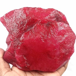 May include: A large, rough-cut ruby gemstone with a deep red color. The raw mineral is held in a hand, showcasing its irregular shape and textured surface. The gemstone is a natural, uncut specimen.