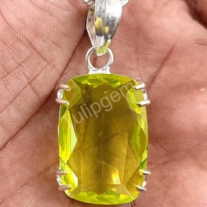 May include: A rectangular, light green gemstone pendant with a silver bail and chain. The gemstone is faceted, and the pendant is held in place by silver prongs. The text "tulipgem" is visible on the gemstone.