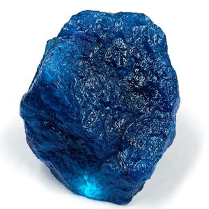 May include: A raw, deep blue gemstone with a textured surface. The stone has a natural, irregular shape and a polished, reflective quality. The colour is a rich, saturated blue, with lighter areas that catch the light.