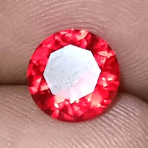 Puede incluir: Una gema roja facetada, de corte redondo, con un centro blanco brillante. La gema tiene un alto grado de claridad y brillo, reflejando la luz. La piedra preciosa es probablemente un rubí o una piedra preciosa similar.