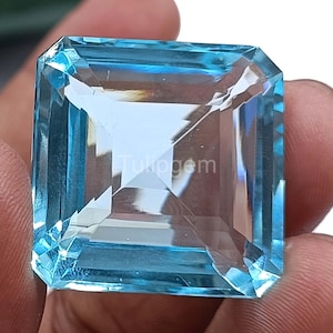 Puede incluir: Una gran gema azul claro, tallada en forma cuadrada, con múltiples facetas. La gema se sostiene entre los dedos, mostrando su claridad y color. El texto "Tulipgem" es visible en la superficie. Una pieza vibrante y llamativa.