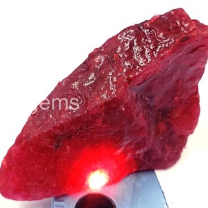 May include: A rough-cut, deep red gemstone with a textured surface. The word "ems" is visible on the stone. The gemstone is illuminated by a bright red light, creating a vibrant display. This natural stone is likely a ruby.