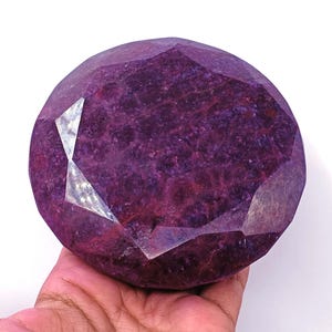 Puede incluir: Una gran gema de rubí facetada con un profundo tono rojo púrpura. La gema tiene múltiples facetas que reflejan la luz, creando un efecto brillante. La piedra se sostiene en una mano, mostrando su tamaño y color.