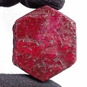 May include: A raw, hexagonal ruby gemstone. The ruby is a deep, vibrant red color with a rough, textured surface. The gemstone is held against a black background.