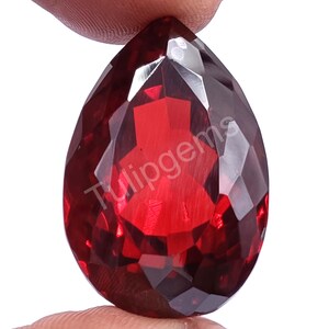 Può includere: Una gemma sfaccettata a forma di goccia, di un colore rosso intenso. La gemma è tenuta tra due dita e presenta il testo "Tulipgems" stampato sulla sua superficie. Il taglio della pietra crea molteplici superfici riflettenti.