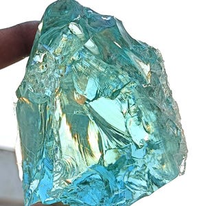 May include: A large, irregularly shaped, translucent blue-green gemstone. The gem has a rough, fractured surface with multiple facets, reflecting light. The colour ranges from light aqua to deeper teal, creating a shimmering effect. The stone is held by a hand.