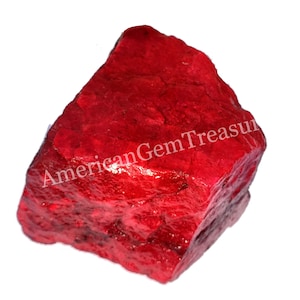 Puede incluir: Una gema roja en bruto, vibrante, con una superficie texturizada. La piedra tiene una forma natural e irregular y muestra un color rojo intenso y rico. El texto "AmericanGemTreasures" es visible. Adecuado para la fabricación de joyas o coleccionar.