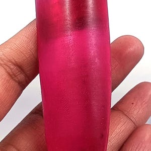 Puede incluir: Una gema rosa vibrante y alargada con una superficie pulida. La gema se sostiene en una mano, mostrando su color y forma. El tono de la piedra varía de un magenta intenso a un rosa más claro en la parte superior, sugiriendo una forma natural.