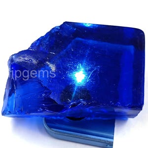 May include: A large, irregularly shaped, deep blue gemstone with a polished surface. The gemstone has a translucent quality, with light reflecting off its facets. The text "ipgems" is visible on the side of the gemstone.