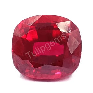 Puede incluir: Una gema de rubí tallada en cojín y facetada. La piedra roja oscura tiene una superficie pulida y el texto "Tulipgems" es visible. La gema está sobre un fondo blanco.