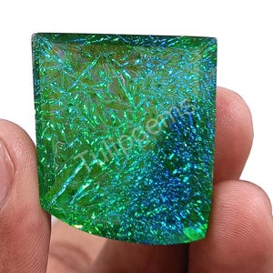 Puede incluir: Una gema iridiscente de forma cuadrada, con un vibrante color verde y azul. La superficie tiene un patrón texturizado y cristalino. La gema se sostiene en una mano sobre un fondo blanco.