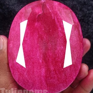 May include: A large, oval-shaped, faceted gemstone in a vibrant pink hue. The gem has a polished surface with internal patterns and white geometric shapes. The text "Tulipgems" is visible at the bottom of the image.