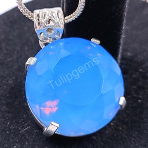 May include: A silver pendant necklace featuring a large, faceted, round, light blue gemstone. The gemstone has a subtle pinkish hue within its depths. The text "Tulipgems" is printed on the gemstone. The necklace chain is also silver.