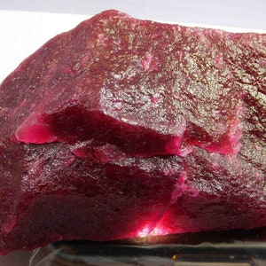 Könnte beinhalten: Ein großer, ungeschliffener Rubin-Edelstein mit tiefroter Farbe. Die Oberfläche hat ein strukturiertes Aussehen mit sichtbaren Kristallstrukturen und Farbintensitätsvariationen. Der Edelstein wird von unten beleuchtet, wodurch ein Leuchteffekt entsteht.