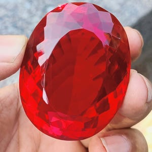 May include: A large, faceted, oval-shaped, deep red gemstone. The gem is held between fingers, showcasing its brilliant, reflective surfaces. The cut of the stone creates numerous facets, enhancing its vibrant color and sparkle. The gemstone appears to be a ruby.