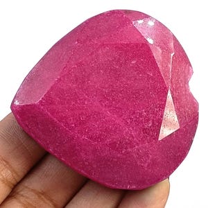 May include: A large, heart-shaped gemstone with a vibrant, saturated magenta-pink colour. The stone features a faceted surface with multiple flat, polished planes that reflect light, creating a textured appearance across its rounded, symmetrical form.