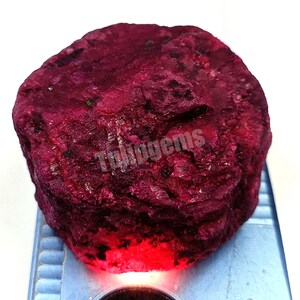 May include: A raw, uncut ruby gemstone in a deep red hue. The surface is textured and uneven, with the word "Tulingems" visible. The gemstone rests on a blue and white surface.