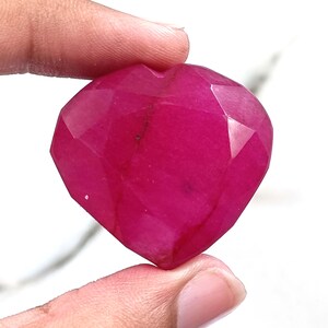 May include: A heart-shaped, magenta-coloured gemstone with multiple facets. The gem is held by fingers, highlighting its polished surfaces and geometric cuts. The gemstone's colour is rich and saturated, reflecting light across its many facets.