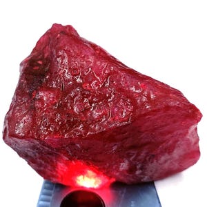 Puede incluir: Una gema de rubí en bruto, sin pulir, de color rojo intenso. La superficie de la piedra tiene una textura con crestas y variaciones visibles. El rubí está iluminado desde abajo, creando un brillo vibrante.