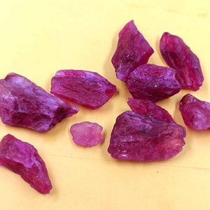May include: A collection of rough, uncut ruby gemstones. The raw rubies display a deep, rich red-pink hue, with visible textures and natural formations. The gemstones are scattered on a light yellow surface.