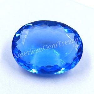 Puede incluir: Una gema ovalada, azul brillante, facetada. La gema tiene una superficie pulida con múltiples facetas que reflejan la luz. El texto "American Gem Treasure" es visible. La gema está sobre un fondo blanco.